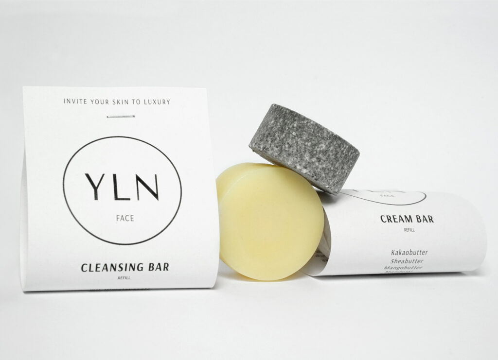 Feste Gesichtsreinigung und Pflege: Der CLEANSING BAR mit Aktivkohle und der CREAM BAR mit pflanzlichen Buttern und Ölen – nebeneinander auf einem Waschbeckenrand, natürliches Licht, minimalistisches Setting.